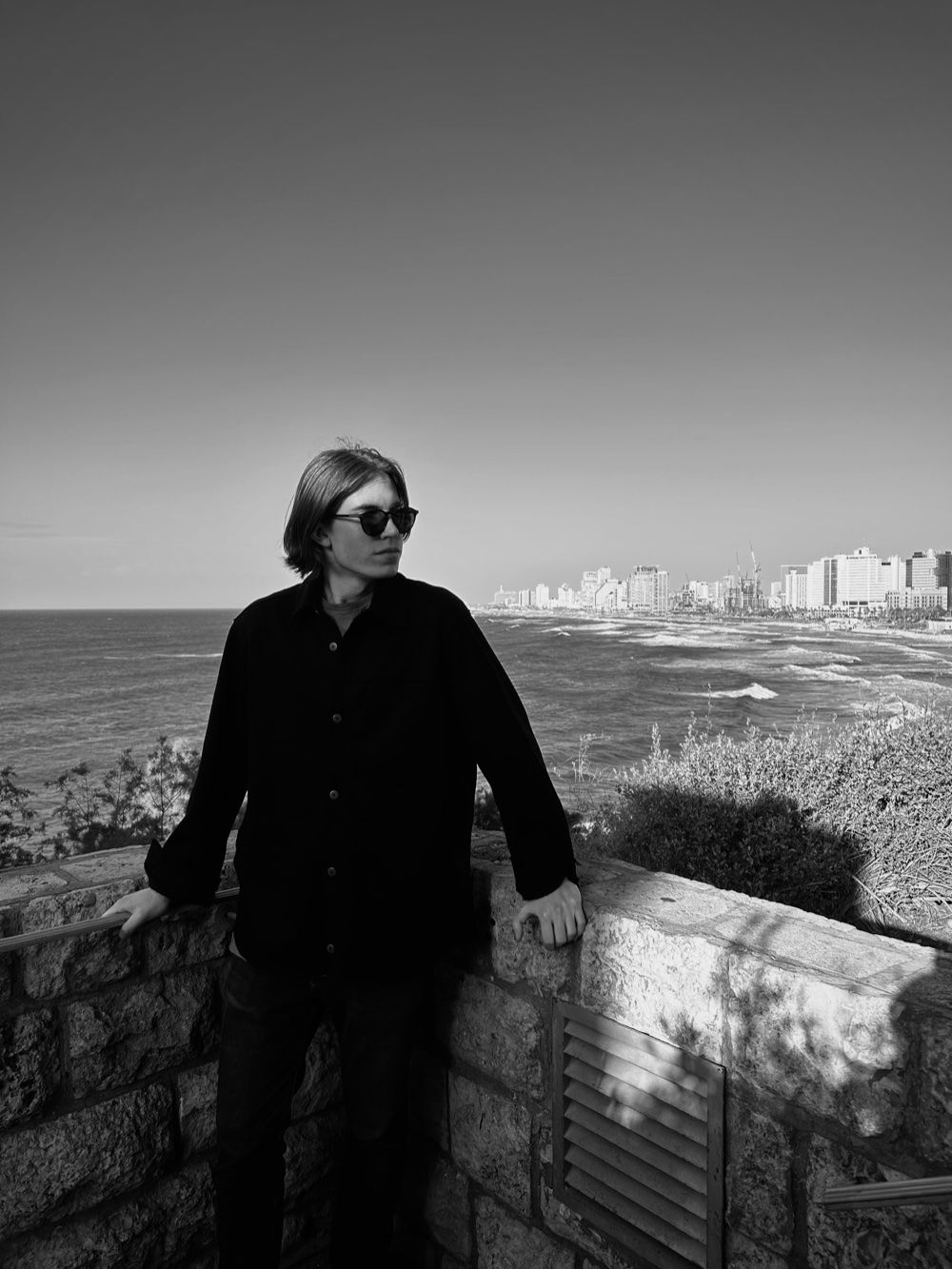 Black-and-white photo of Denis Maksimov leaning on a stone wall by the sea